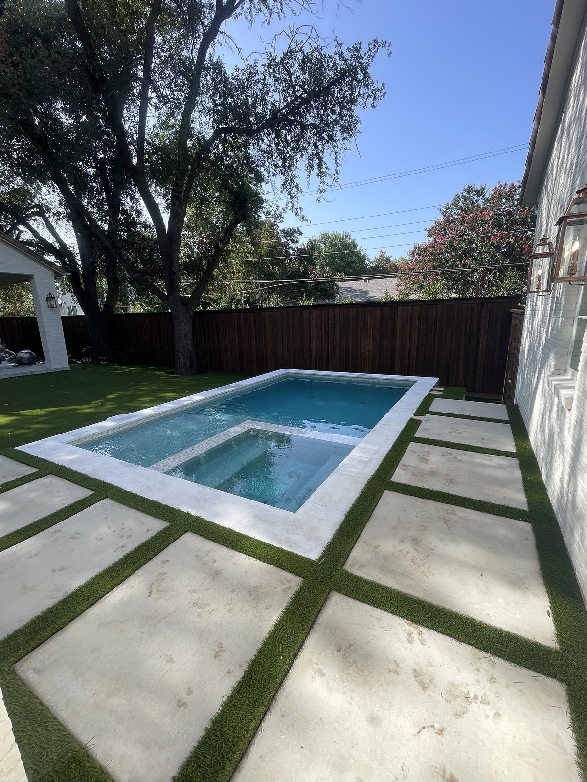 Full backyard view: rectangular pool with built-in spa, white stone coping, concrete pavers with turf joints and artificial lawn under mature trees — completed by Infinity Group DFW in Dallas, Texas